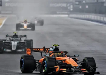 Mobil McLaren Formula 1 berwarna oranye melaju di lintasan basah, diikuti beberapa mobil lain
