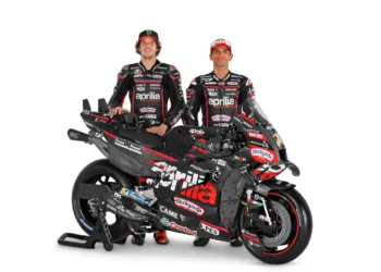 Dua pembalap berseragam Aprilia Racing berdiri di belakang motor balap RS-GP26 dengan latar putih