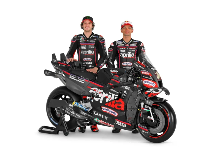 Dua pembalap berseragam Aprilia Racing berdiri di belakang motor balap RS-GP26 dengan latar putih
