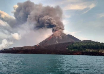 Ilustrasi gunung berapi dengan kepulan abu tebal di atas kawah, terlihat dari perairan