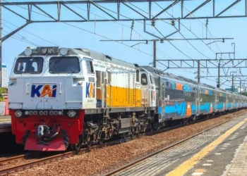 Kereta penumpang dengan lokomotif KAI berwarna putih dan kuning berada di jalur stasiun, dengan rangkaian gerbong biru di belakangnya