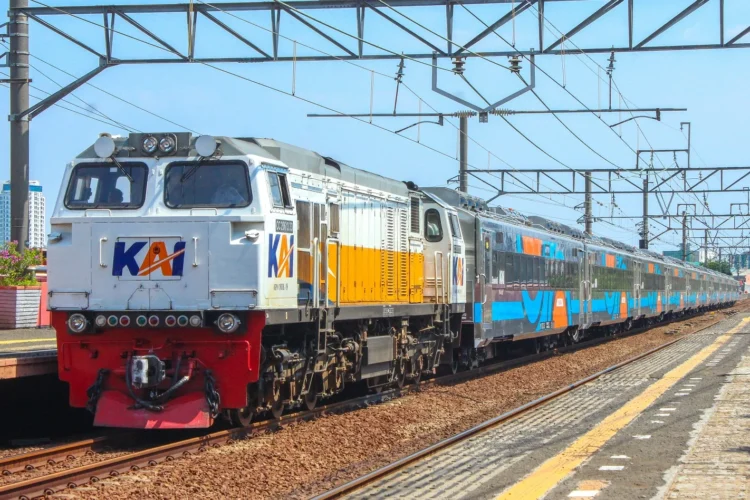Kereta penumpang dengan lokomotif KAI berwarna putih dan kuning berada di jalur stasiun, dengan rangkaian gerbong biru di belakangnya