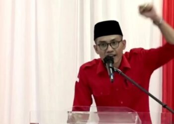 Pria berkemeja merah dan berpeci hitam berbicara melalui mikrofon sambil mengangkat tangan di depan podium
