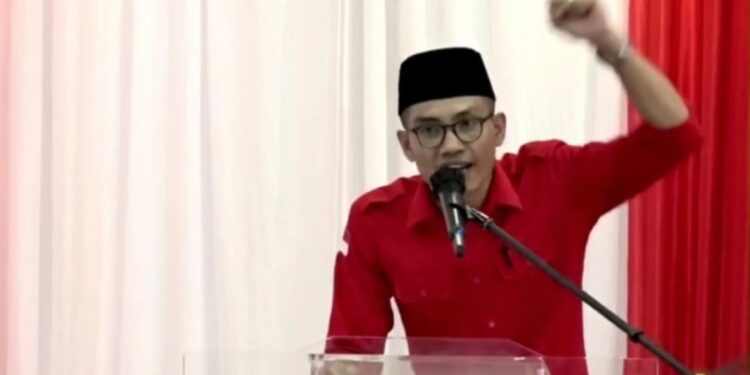 Pria berkemeja merah dan berpeci hitam berbicara melalui mikrofon sambil mengangkat tangan di depan podium
