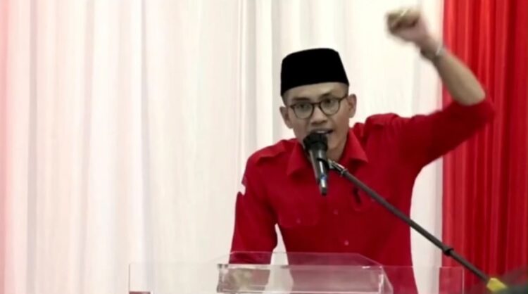 Pria berkemeja merah dan berpeci hitam berbicara melalui mikrofon sambil mengangkat tangan di depan podium