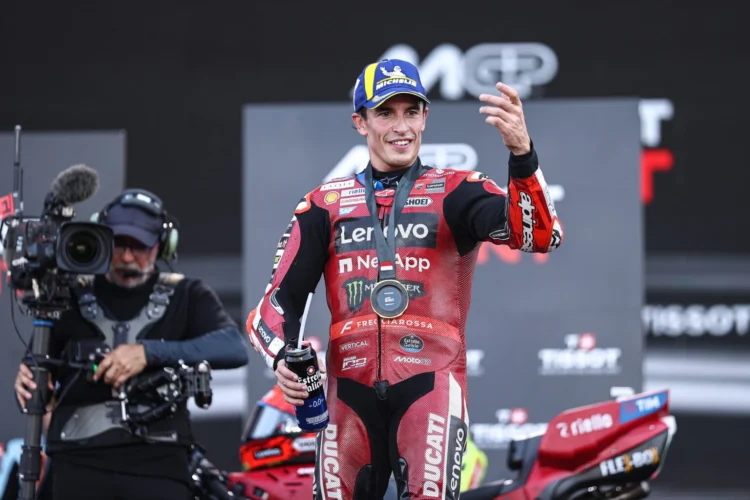 Pembalap Ducati Lenovo Team mengenakan baju balap merah dan topi biru sambil melambaikan tangan di depan kamera televisi
