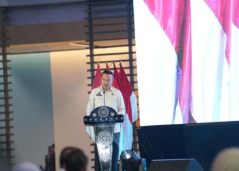 Direktur Jenderal Imigrasi berbicara di podium pada sebuah acara resmi dengan bendera Indonesia di belakangnya
