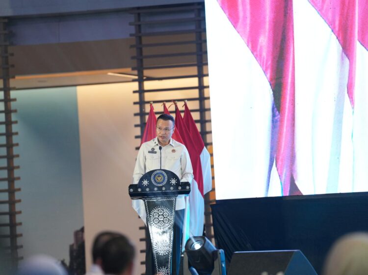 Direktur Jenderal Imigrasi berbicara di podium pada sebuah acara resmi dengan bendera Indonesia di belakangnya