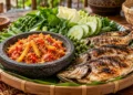 Ilustrasi ikan bakar disajikan bersama sambal seruit merah, lalapan, dan nasi dalam tampah