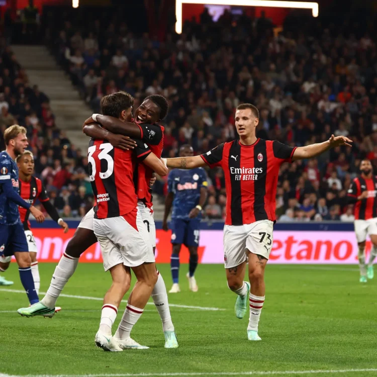 Para pemain AC Milan berseragam merah dan hitam saling berpelukan merayakan gol, dengan pemain lawan berbaju biru di lapangan