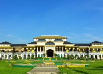 Tampak depan Istana Maimun berwarna kuning dan putih dengan atap gelap, taman hijau, dan jalan setapak di halaman