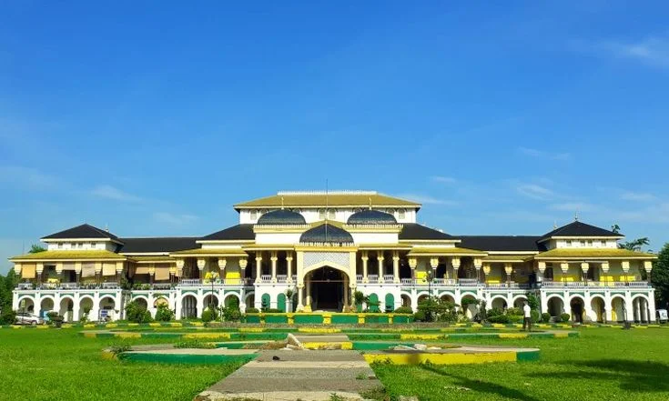 Tampak depan Istana Maimun berwarna kuning dan putih dengan atap gelap, taman hijau, dan jalan setapak di halaman