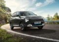 Kia Carens EV berwarna abu-abu gelap melaju di jalan berkelok dengan pepohonan dan pagar pembatas di sisi jalan.