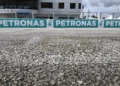 Foto jarak dekat permukaan aspal Sirkuit Sepang, dengan papan Petronas dan bangunan sirkuit di belakang