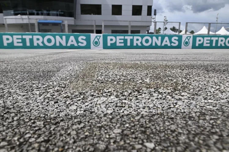 Foto jarak dekat permukaan aspal Sirkuit Sepang, dengan papan Petronas dan bangunan sirkuit di belakang
