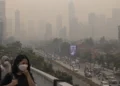Ilustrasi warga mengenakan masker di jembatan penyeberangan dengan gedung perkotaan dan jalan raya yang tampak berkabut di belakangnya