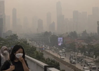 Ilustrasi warga mengenakan masker di jembatan penyeberangan dengan gedung perkotaan dan jalan raya yang tampak berkabut di belakangnya