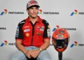 Marc Marquez mengenakan kemeja tim Ducati berwarna merah, duduk di sebelah helm balap dengan latar logo Pertamina