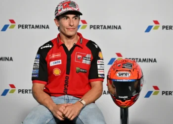 Marc Marquez mengenakan kemeja tim Ducati berwarna merah, duduk di sebelah helm balap dengan latar logo Pertamina