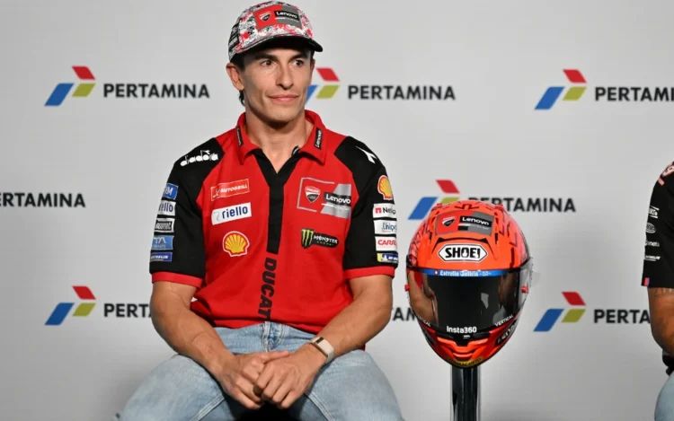 Marc Marquez mengenakan kemeja tim Ducati berwarna merah, duduk di sebelah helm balap dengan latar logo Pertamina