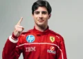 Seorang pembalap muda memakai seragam balap merah berlambang Ferrari dan mengangkat satu jari dengan latar polos