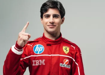 Seorang pembalap muda memakai seragam balap merah berlambang Ferrari dan mengangkat satu jari dengan latar polos