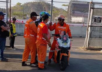 Beberapa petugas berseragam oranye berdiri di dekat skuter operasional di samping pagar area sirkuit