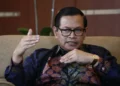 Pramono Anung mengenakan kacamata dan kemeja batik sambil berbicara dengan tangan terangkat saat duduk di kursi