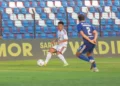 Pemain berseragam putih menguasai bola di depan pemain berseragam biru, dengan tribun stadion di belakang