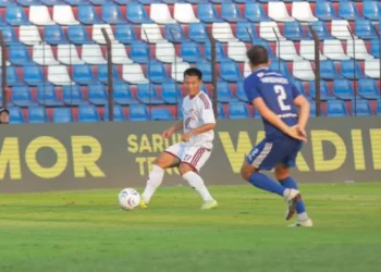 Pemain berseragam putih menguasai bola di depan pemain berseragam biru, dengan tribun stadion di belakang