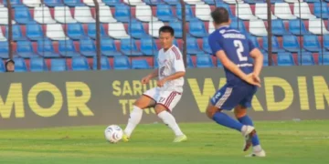 Pemain berseragam putih menguasai bola di depan pemain berseragam biru, dengan tribun stadion di belakang