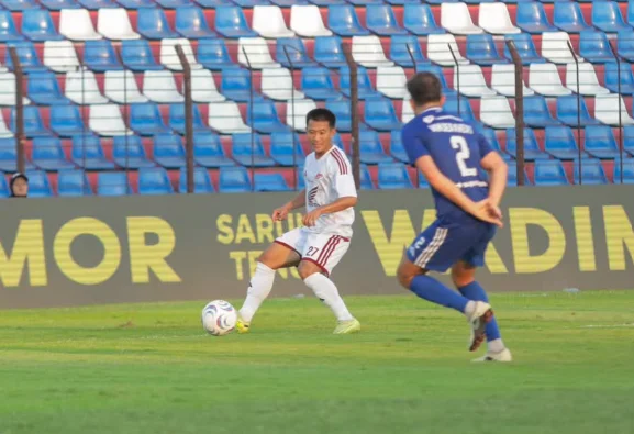 Pemain berseragam putih menguasai bola di depan pemain berseragam biru, dengan tribun stadion di belakang