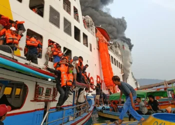 Penumpang berpelampung oranye dipindahkan dari kapal penyeberangan berasap ke perahu-perahu kecil di laut