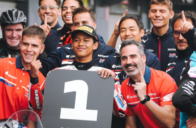 Veda Ega Pratama mengenakan seragam Honda Team Asia dan berpose bersama anggota tim di belakang papan nomor satu setelah meraih pole position Moto3.