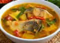 Potongan ikan patin dalam kuah kuning kemerahan dengan cabai, sayuran, dan daun hijau di mangkuk putih