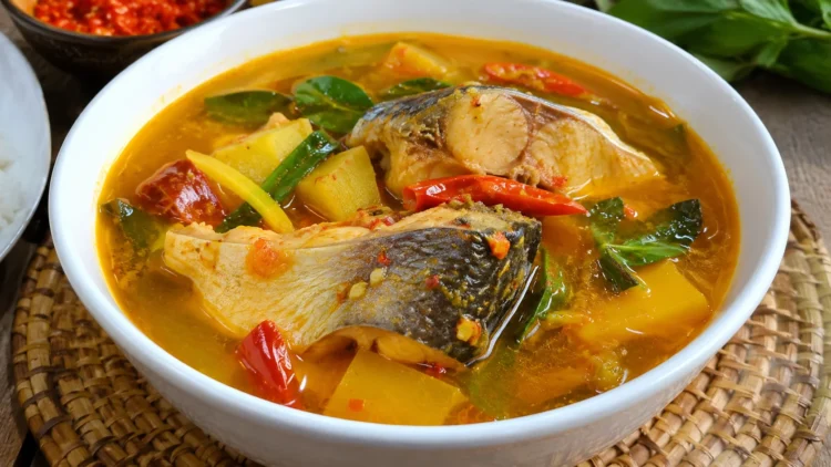 Potongan ikan patin dalam kuah kuning kemerahan dengan cabai, sayuran, dan daun hijau di mangkuk putih