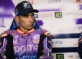 Toprak Razgatlioglu mengenakan baju balap Prima Pramac Yamaha berwarna ungu dan oranye serta topi hitam saat berada di area tim.