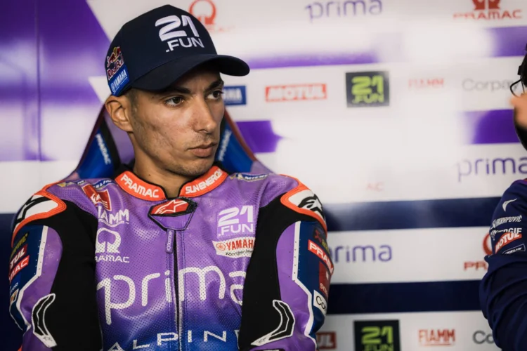 Toprak Razgatlioglu mengenakan baju balap Prima Pramac Yamaha berwarna ungu dan oranye serta topi hitam saat berada di area tim.