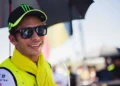 Valentino Rossi tersenyum dengan topi hitam, kacamata hitam, dan syal berwarna kuning