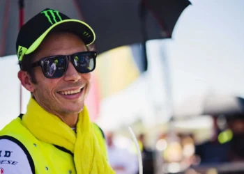 Valentino Rossi tersenyum dengan topi hitam, kacamata hitam, dan syal berwarna kuning