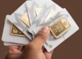 Tangan memegang beberapa emas batangan Logam Mulia dalam kemasan putih dengan latar belakang cokelat