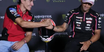 Marc Marquez mengenakan kemeja Ducati merah berjabat tangan dengan Jorge Martin berbaju tim Aprilia hitam, dengan helm di antara mereka