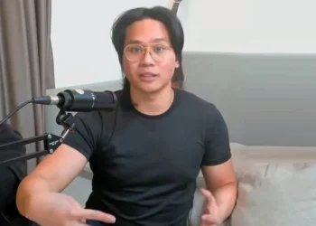 Pria berkacamata dan mengenakan kaus hitam berbicara di depan mikrofon dalam sebuah rekaman video