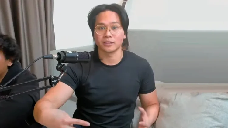 Pria berkacamata dan mengenakan kaus hitam berbicara di depan mikrofon dalam sebuah rekaman video