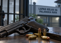 Ilustrasi pistol, peluru, dan magasin di meja dengan gambaran gerbang lapas serta petugas di latar belakang