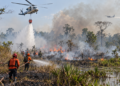Ilustrasi helikopter menyiram air ke lahan terbakar sementara petugas memadamkan api dan pesawat melintas di kejauhan