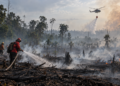 Ilustrasi petugas menyiram area bekas kebakaran hutan yang masih berasap, dengan helikopter pemadam di kejauhan