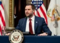 Wakil Presiden AS JD Vance mengenakan jas biru dan dasi merah berbicara di depan mikrofon dengan bendera Amerika Serikat di belakangnya