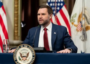 Wakil Presiden AS JD Vance mengenakan jas biru dan dasi merah berbicara di depan mikrofon dengan bendera Amerika Serikat di belakangnya