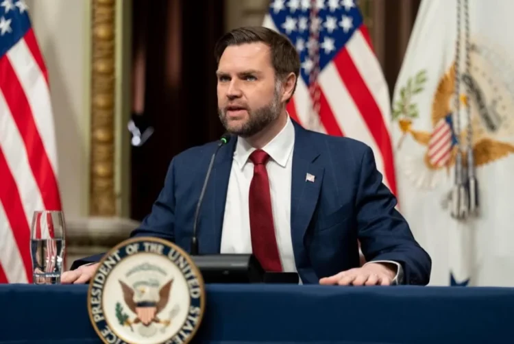 Wakil Presiden AS JD Vance mengenakan jas biru dan dasi merah berbicara di depan mikrofon dengan bendera Amerika Serikat di belakangnya